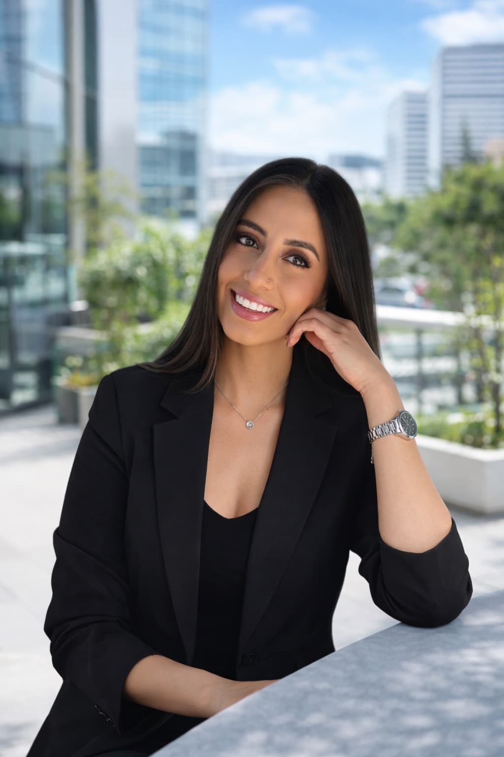 Professional woman with long dark hair smiling in a black blazer against a city backdrop.