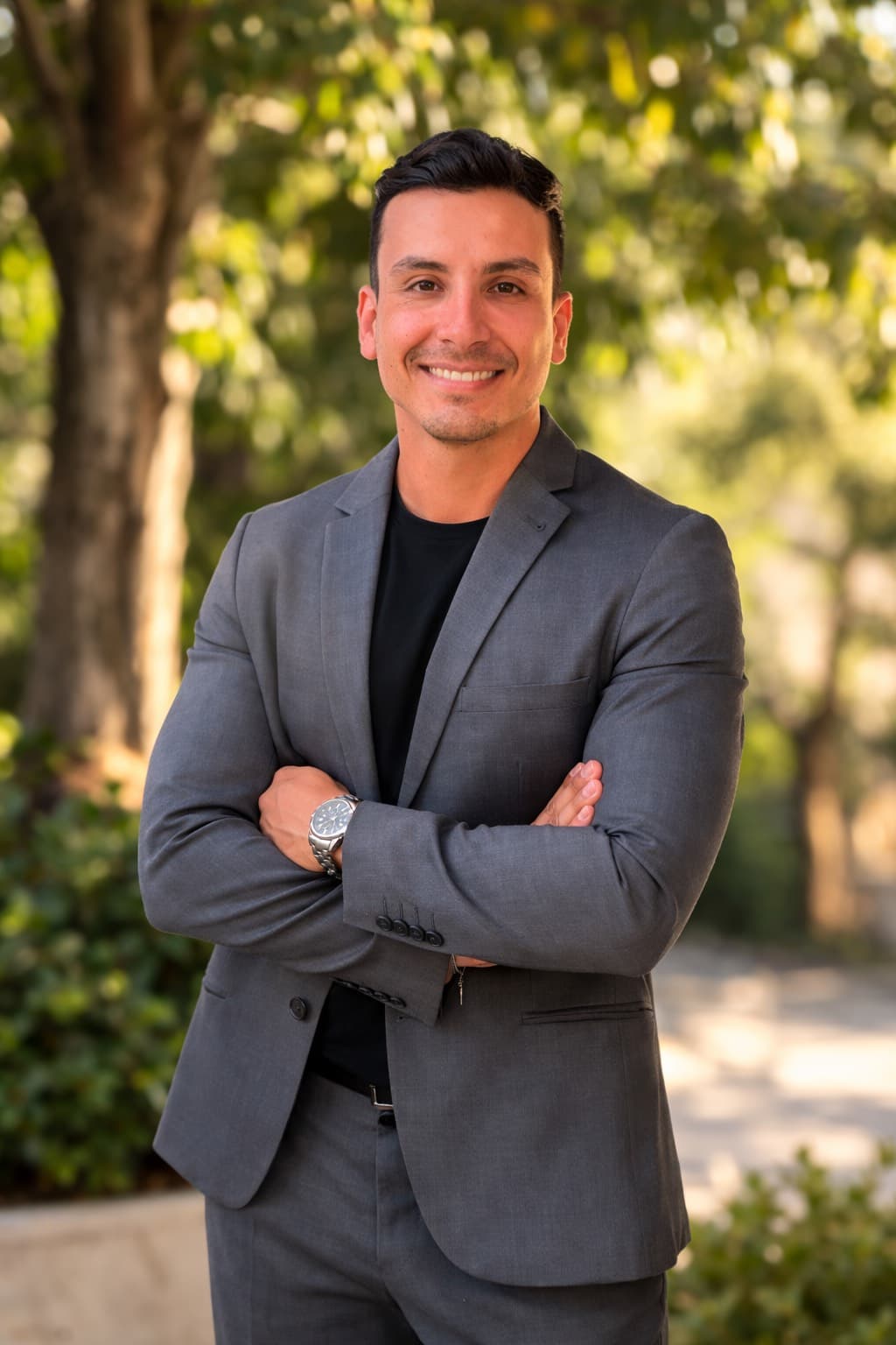 Smiling man in a grey suit jacket and black shirt with arms crossed outdoors.