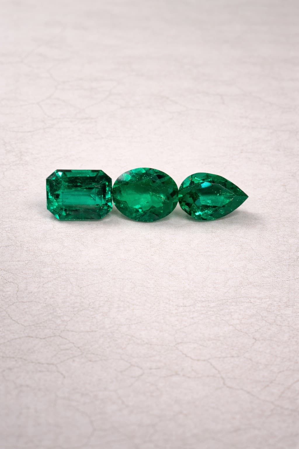 Three vibrant green emeralds in emerald, oval, and pear cuts on a textured white surface.