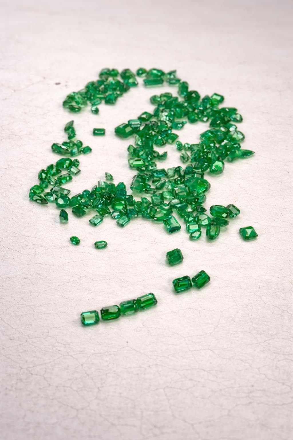 Numerous faceted green emeralds in various cuts scattered across a light, textured stone surface.
